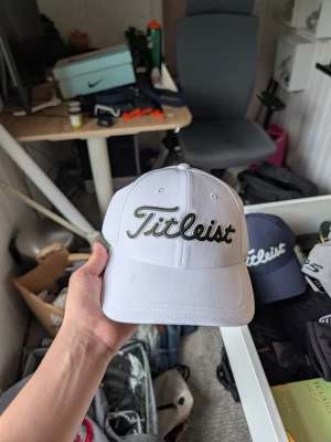 Vit keps från Titleist - Snygg vit keps från Titleist med broderad logga framtill i svart och grått. Kepsen har böjd skärm och justerbar rem baktill. Tillverkad i ett lätt och ventilerande material, perfekt för golf eller vardagsstil. Inbyggd magnet i skärmen för marker.