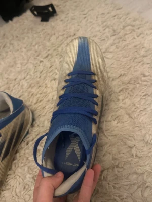 Adidas X blå/vita fotbollsskor - Säljer ett par Adidas X fotbollsskor i blått och vitt med klassiska tre svarta ränder på sidan. Skorna har snörning och ovandel i syntetmaterial för bra passform och lätt känsla. Perfekta för snabba spelare som vill ha kontroll och fart på planen.