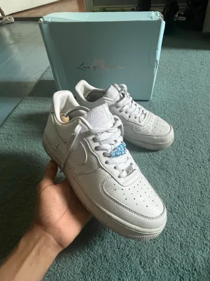 Nike Air Force 1 x Love You Forever - Nike Air Force 1 x Love You Forever i helvitt läder med klassisk siluett och perforerade detaljer på tån. Swoosh-logga på sidorna och blå detalj på snörningen. Boxen har Love You Forever-tryck. Perfekta sneakers för dig som gillar stilrena och ikoniska skor.detta är ett samarbete mellan nike och nocta