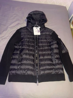 moncler cardigan - Snygg svart pufferjacka från Moncler med quiltad front och huva. Ärmar och rygg är i ribbstickad bomull vilket ger en cool kontrast. Jackan har dragkedja framtill, två fickor med knapp och Moncler-logga på ärmen. Perfekt för kalla vinterdagar.
