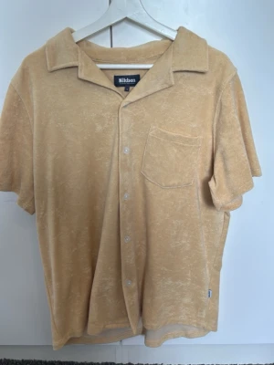 Beige frottéskjorta från Nikben - Säljer en beige skjorta från Nikben i mjuk frotté med kort ärm, klassisk krage och bröstficka. Perfekt för en avslappnad och trendig look. Skjortan har knäppning framtill och en ledig passform.
