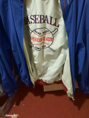 Gant vindjacka med baseball-tryck - Retro vindjacka från Gant med stort baseball-tryck på ryggen och framsidan. Jackan är vit med blå ärmar och röda detaljer vid krage och midja. Oversized passform och elastiska muddar. Perfekt för dig som gillar sportig och amerikansk stil.
