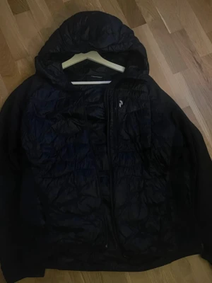 Svart pufferjacka från Peak Performance - Snygg svart pufferjacka från Peak Performance med huva och dragkedja framtill. Jackan har quiltad design och är tillverkad i ett glansigt, lätt material som håller värmen. Perfekt för kalla vinterdagar och har en diskret logga på bröstet. De står XXL som storlek men de är i junior storlek och de är L i herr storlek 