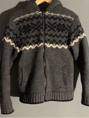 Grå stickad hoodie med mönster - Mysig grå stickad hoodie med dragkedja och klassiskt nordiskt mönster i svart och vitt över bröst och axlar. Perfekt för för sommarkvällar! Skick 10/10 knappt använd. Storlek M men sitter som S