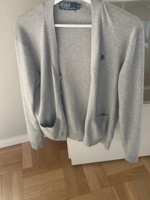 Grå cardigan från Polo Ralph Lauren - Snygg grå cardigan från Polo Ralph Lauren med två fickor framtill och klassisk broderad logga på bröstet. Långärmad och med knäppning framtill. Perfekt att slänga över en t-shirt för en chill look.