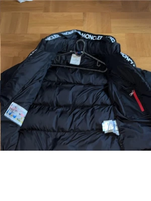 Svart Moncler dunjacka med logga - Snygg svart dunjacka från Moncler med stora vita loggor längs dragkedjan och huvan. Jackan har en puffig design, röda detaljer på fickorna och är tillverkad i glansigt material. Perfekt för kalla vinterdagar.