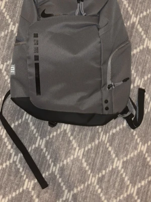 Grå Nike Elite ryggsäck - Snygg grå ryggsäck från Nike Elite med svarta detaljer och vit logga på axelremmarna. Ryggsäcken har flera fack, dragkedjor och justerbara remmar. Perfekt för skolan eller träningen. Materialet är slitstark polyester.