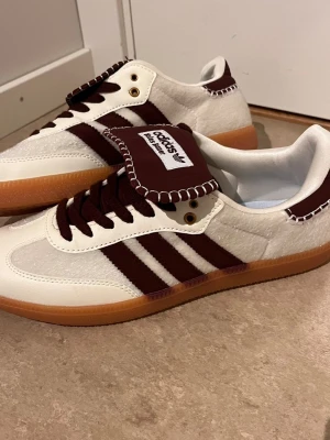 Adidas Samba OG vit och vinröd - Adidas Samba OG sneakers i vitt och vinrött med klassiska tre ränder på sidan, brun gummisula och detaljer i mocka och läder. Tungan har Adidas-logga och kontrastsömmar. Snygg retrostil som sticker ut.