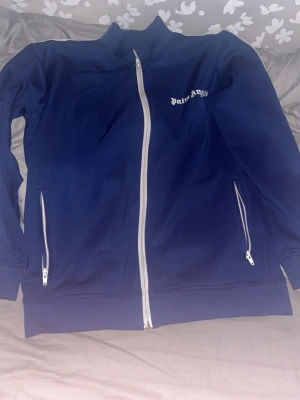 Palm angels  - Blå track jacket från Palm Angels med vita dragkedjor och vita ränder längs ärmarna. Jackan har ståkrage, två fickor med dragkedja och logga på bröstet. Perfekt för en sportig och trendig stil.