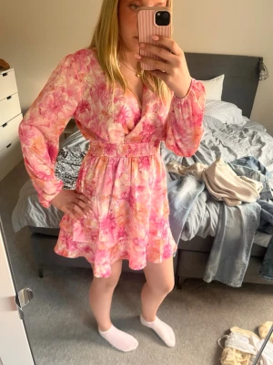 Kort sommarklänning - Söt kort klänning med blommigt mönster i rosa och orange toner. Klänningen har v-ringning, långa ärmar och smock i midjan för en bekväm passform. Volangdetalj nertill ger en extra feminin touch. Perfekt för vår och sommar!