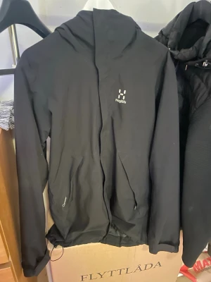 Svart vindjacka från Haglöfs - Snygg svart vindjacka från Haglöfs med dragkedja framtill och två fickor. Jackan har en minimalistisk design med diskret logga på bröstet och huva. Perfekt för blåsiga dagar och enkel att matcha med olika . Lite trasig bara 