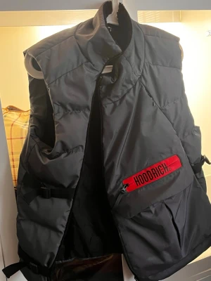Svart dunväst från Hoodrich - Snygg svart dunväst från Hoodrich med hög krage och cool röd detalj med logga på bröstet. Västen har dragkedja och praktiska fickor framtill. Perfekt för lager-på-lager och streetwear-stil.