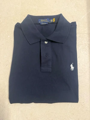 Ralph Lauren piké  - Klassisk marinblå pikétröja från Polo Ralph Lauren med vit broderad logga på bröstet. Tröjan har krage, korta ärmar och knäppning framtill. Perfekt för en stilren och avslappnad look.