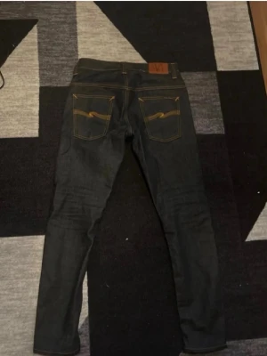 Mörkblå jeans från Nudie Jeans - Säljer ett par mörkblå slim fit jeans från Nudie Jeans med klassiska kontrastsömmar och snygga bakfickor med den ikoniska Nudie-bågen. Jeansen har fem fickor och är tillverkade i kraftig denim, perfekta för en clean och stilren look. W29 L32