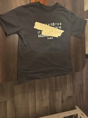 Svart Maison Margiela t-shirt - Svart t-shirt från Maison Margiela med ikoniskt tryck framtill i vitt och beige. Klassisk passform med rund hals och korta ärmar. Märkesetikett syns på ryggen. Perfekt för dig som gillar streetwear och designerplagg. Den är i modellen lite oversized som på senare tid har blivit populärt