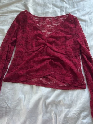 Vinröd spetsblus med v-ringning - Vinröd långärmad blus i spets med v-ringning och snygg rynkdetalj framtill. Blusen är genombruten och har en feminin, croppad passform som passar perfekt till jeans eller kjol.