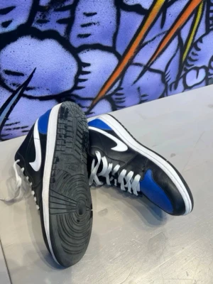 Nike Air Jordan 1 Low blå/svart/vit - Snygga Nike Air Jordan 1 Low sneakers i blått, svart och vitt läder med klassisk vit swoosh på sidorna. Skorna har vita skosnören och den ikoniska Air Jordan-loggan på hälen. Perfekta för dig som vill sticka ut med en streetwear-look.