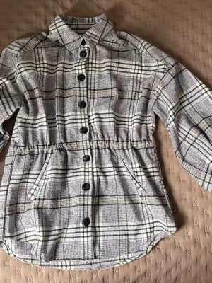 Rutig overshirt jacka med knappar - Snygg overshirt i grått, beige och svart rutmönster med knappar framtill och två snedställda fickor. Overshirten har krage och resår i midjan för en schysst passform. Perfekt lager-på-lager-plagg för en trendig look.