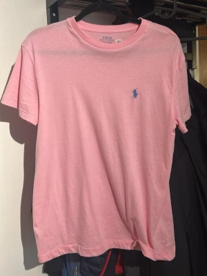 Rosa t-shirt från Polo Ralph Lauren - Snygg rosa t-shirt från Polo Ralph Lauren med klassisk passform och blå logga broderad på bröstet. Använd 1 gång. Tillverkad i mjuk bomull och har korta ärmar. Perfekt för en avslappnad och stilren look.