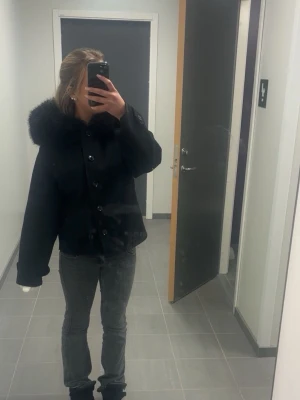 Svart jacka med fluffig luva - Säljer en svart jacka med stor fluffig luva . Jackan har knäppning framtill med stora knappar. Perfekt för kalla vinterdagar men speciellt på hösten! Också bra när det regnar
