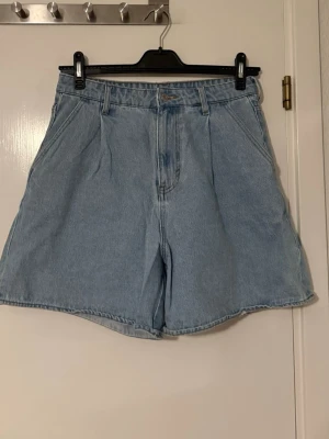 Ljusblåa high waist denimshorts &Denim H&M - Säljer ett par ljusblåa denimshorts från &Denim med hög midja och bred passform. Klassisk femficksmodell med dragkedja och knapp framtill. Perfekta för en chill och avslappnad stil under varmare dagar.