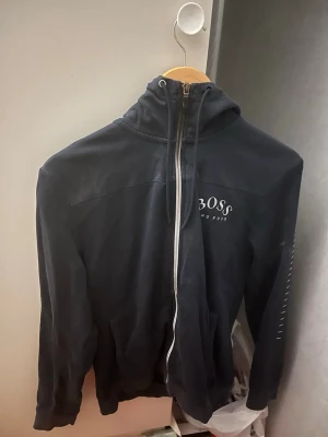 Svart hoodie från BOSS med dragkedja - Snygg svart hoodie från BOSS med vit dragkedja och logga på bröstet. Modellen har huva och långa ärmar, samt diskreta detaljer på ena ärmen. Perfekt för en avslappnad och stilren look. Priser kan diskuteras 