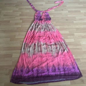 Lång färgglad klänning med halterneck - Snygg långklänning med halterneck och v-ringning. Klänningen har ett färgstarkt mönster i rosa, lila och beige toner och är tillverkad i ett lätt och luftigt material. Perfekt för varma dagar och somriga tillfällen.