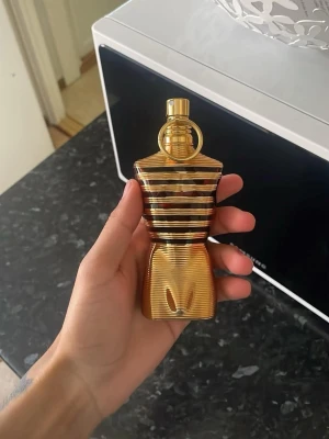 Jean Paul Gaultier Le Male Le Parfum - Säljer en ikonisk parfym från Jean Paul Gaultier, Le Male Le Parfum. Flaskan är formad som en manlig torso i guld med svarta och bronsfärgade ränder. Flaskan är tillverkad i glas och metall och har en unik, lyxig design som sticker ut i parfymhyllan.