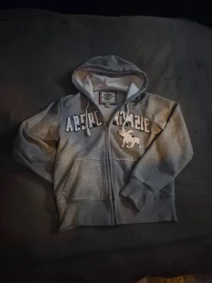 Grå hoodie från Abercrombie & Fitch - Grå hoodie med dragkedja från Abercrombie & Fitch. Stor logga och broderad älg på bröstet, huva med snörning och två fickor framtill. Mjuk insida och ribbade muddar. Perfekt för en avslappnad stil.