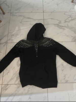 Svart hoodie med vingar från Marcelo Burlon - Snygg svart hoodie från Marcelo Burlon med ikoniskt vingtryck i vitt och grått över axlar och bröst. Klassisk huva och känguruficka framtill. Perfekt för dig som vill sticka ut med en unik design.