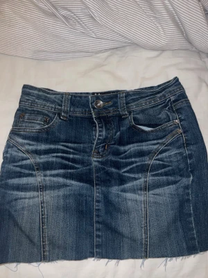 Blå jeanskjol lågmidjad  - Snygg blå lågmidjad jeanskjol i klassisk denim med rå, fransig kant nertill. Kjolen har fem fickor, bälteshällor och dragkedja med knapp framtill. Perfekt för en avslappnad och trendig look.