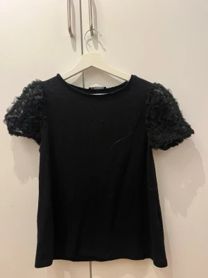 Svart topp med puffärmar - Svart topp med rund halsringning och korta puffärmar i ett fluffigt, texturerat material. Ärmarna ger en cool och trendig look medan resten av toppen är slät och enkel. Perfekt för dig som vill sticka ut lite extra.