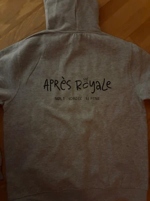 Grå hoodie Après Royale - Säljer en grå hoodie från Nolt Nordic Alpine med trycket 'Après Royale' på ryggen. Hoodien har huva och är tillverkad i mjukt material, perfekt för chill dagar. Enkel och stilren design med svart text och liten krona ovanför O:et. Tröja är tryck fel och därför priset!