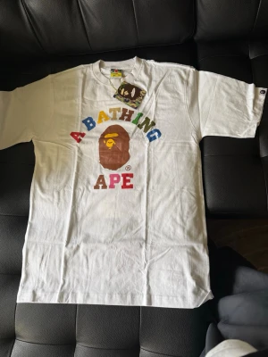 Vit A Bathing Ape t-shirt med tryck - Vit t-shirt från A Bathing Ape med färgglatt tryck av texten 'A BATHING APE' och det klassiska apansikte-loggan på bröstet. T-shirten har rund hals och korta ärmar. Materialet är mjuk bomull och passformen är normal.