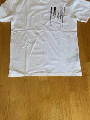 Vit AMIRI t-shirt med färgstänk - Säljer en vit t-shirt från AMIRI med tryck av loggan och färgstänk i flera färger på både fram- och baksidan. T-shirten har klassisk passform och är kortärmad, tillverkad i mjuk bomull. Perfekt för dig som gillar streetwear med en unik twist.