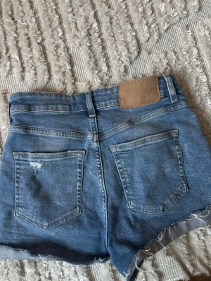 Blå jeansshorts från H&M - Snygga blå jeansshorts från H&M med råa kanter och klassisk femficksdesign. Shortsen har hög midja och en liten slitning på ena bakfickan för en cool, avslappnad look. Perfekta för varma dagar och enkel att matcha med allt.