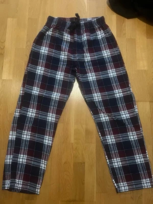Rutiga pyjamasbyxor i flanell - Mysiga pyjamasbyxor i mjuk flanell med rutigt mönster i marinblått, vinrött och vitt. Byxorna har resår i midjan och snörning för bästa passform. Perfekta för chillkvällar hemma eller när du vill vara extra bekväm.