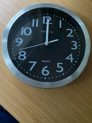 Väggklocka Moretime svart och silver - Stilren väggklocka från Moretime med svart urtavla, tydliga vita siffror och visare. Klockan har en rund form och en ram i silverfärgad metall. Drivs med quartz-urverk och passar perfekt i kök, vardagsrum eller kontor.