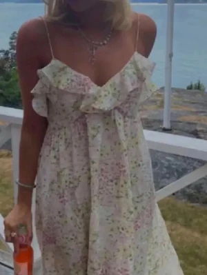 Blommig långklänning med volang - Romantisk långklänning med smala axelband och volangdetalj vid urringningen. Klänningen är vit med blommigt mönster i rosa och grönt, har en luftig passform och är perfekt för varma dagar.