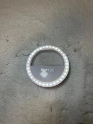 Selfie-ringlampa från Catly - Rund selfie-ringlampa i vit plast från Catly. Lampan har LED-ljus runt hela kanten och ett klämfäste som gör den enkel att fästa på mobilen eller spegeln. Perfekt för att få jämnt ljus till sminkning eller selfies.