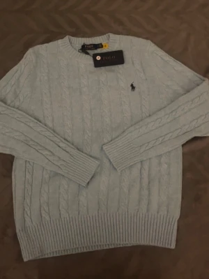 Ljusblå kabelstickad tröja Polo Ralph Lauren - Snygg ljusblå kabelstickad tröja från Polo Ralph Lauren med rund halsringning och klassisk broderad logga på bröstet. Tröjan har långa ärmar och ribbstickade muddar vid ärmslut och nederkant. Perfekt för en stilren look.