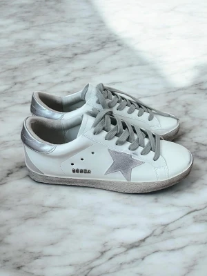 Golden Goose vita sneakers med silverdetaljer - Snygga vita sneakers från Golden Goose med silverfärgad stjärna på sidan, silvriga detaljer vid hälen och grå skosnören. Skorna har en klassisk låg modell och är tillverkade i läder med slitna detaljer på sulan för en cool look.