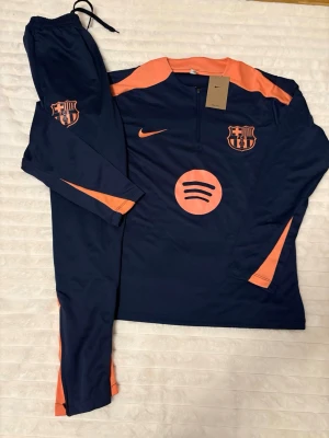 Barça träningsset Nike mörkblå/orange - Säljer ett FC Barcelona träningsset från Nike i mörkblått med orange detaljer. Setet består av långärmad tröja med halv dragkedja och matchande byxor. Både tröja och byxor har klubbmärke och Nike-logga, samt Spotify-logga på bröstet. Perfekt för fotbollsträning.
