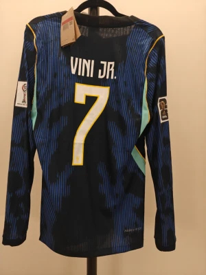 Spelar Version Brasilien Vini Jr. långärmad matchtröja - Snygg spelar Version långärmad fotbollströja från Nike för Brasilien med Vini Jr. och nummer 7 på ryggen. Tröjan är blå med svarta och gula detaljer, officiella FIFA och CBF-emblem samt Aeroready-material för bra ventilation. Perfekt för fotbollsfans!