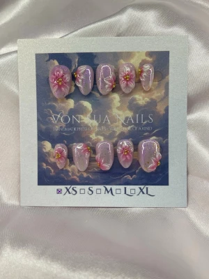 SakuraDrip (XS);ExtraShortAlmond - Handgjorda press-on naglar i rosa/lila med glittrig finish och handgjorda 3D-blommor i rosa och vitt med gulddetaljer. Naglarna har en extra kort mandel form och kommer i XS: 14 mm, 11 mm, 11,5 mm, 10 mm, 8 mm . Perfekt för dig som vill ha en unik och kreativ nageldesign.