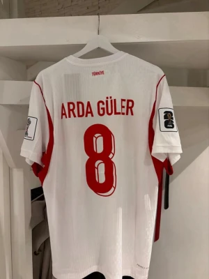 Turkiet matchtröja Arda Güler Nike - Säljer en vit och röd matchtröja från Nike med Arda Güler och nummer 8 på ryggen. Tröjan har Turkiets flagga på bröstet, röda detaljer och FIFA-märke på ärmarna. Perfekt för dig som älskar fotboll och vill stötta Turkiet på match! tröjan är i Player fit så dene slim fit// MOTSVARAR XL/L