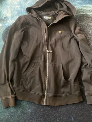 Svart hoodie från Lyle & Scott - Svart hoodie med dragkedja från Lyle & Scott. Klassisk modell med huva, två fickor framtill och gul logga på bröstet. Tillverkad i mjukt bomullsmaterial, perfekt för en avslappnad stil.