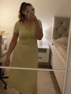 Gul klänning med snörning i midja - Söt gul klänning med kort ärm och snörning vid bysten. Klänningen har en lätt utställd form och fyrkantig ringning, vilket ger en feminin och trendig look. Perfekt för varma dagar och somriga tillfällen.