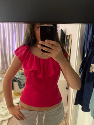 Rosa volangtopp med kort ärm - Snygg rosa topp med volangdetalj längs ringningen och korta ärmar. Modellen är figurnära och toppen har en feminin och trendig look. Perfekt att styla med jeans eller kjol för en cool vardagsstil.