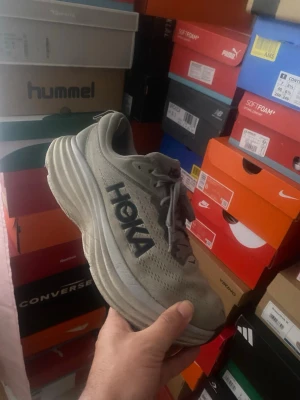 Gråa Hoka bondi 8  - Säljer ett par gråa Hoka sneakers med riktigt chunky och hög sula. Skorna har ventilerande meshmaterial och klassisk snörning. Hoka-loggan syns tydligt på sidan. Perfekta för dig som gillar sportig och modern stil.
